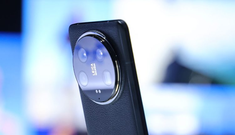 Close-up of a smartphone featuring a prominent circular Leica-branded camera module with multiple lenses, mounted on a sleek, leather-textured back against a blurred background
