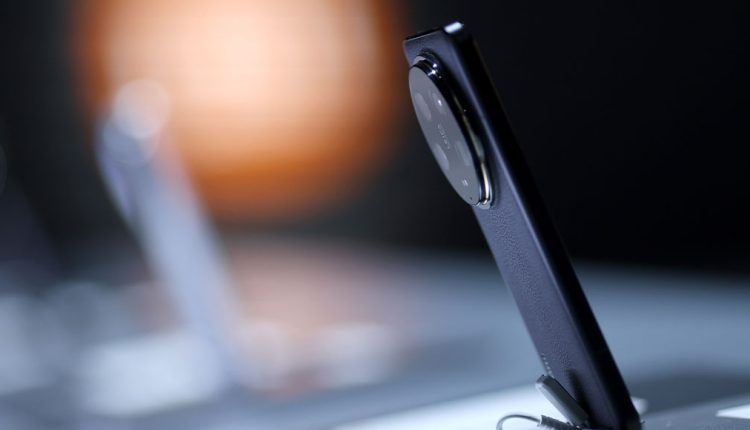 Side view of a modern smartphone on display, featuring a large circular rear camera module and a sleek leather-textured back, positioned upright on a showcase stand with blurred background lighting