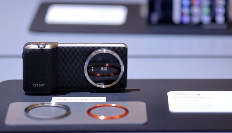 Xiaomi smartphone with a Leica-branded circular camera module showcased on a display table, designed to resemble a compact digital camera, with additional lens accessories placed in front