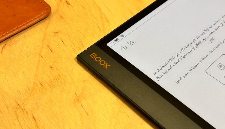 A close-up, angled shot focusing on the left edge of a Boox Note Air3 C e-reader tablet, which features the orange "BOOX" logo. The screen displays text in Arabic. The device is resting on a wooden surface, with a corner of a tan leather object visible in the upper left.