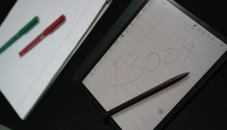 An overhead, angled shot showing a Boox Note Air3 C e-reader tablet with its screen displaying a digital notepad with grid lines and the word "BOOX" handwritten on it. A black stylus pen rests on the screen. To the left, a physical notebook with lined paper is visible, along with two colorful pens (green and red).