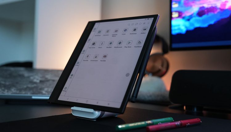 A Boox Note Air3 C e-reader tablet stands upright on a white stand on a dark desk. Its screen shows a grid of application icons with labels like "Notebook," "Calendar," "Gallery," and "YouTube." A black stylus pen is magnetically attached to the side of the tablet. In the foreground, two colorful pens (green and red) are visible. The background shows a dimly lit room with a monitor displaying colorful abstract art and other desk items.