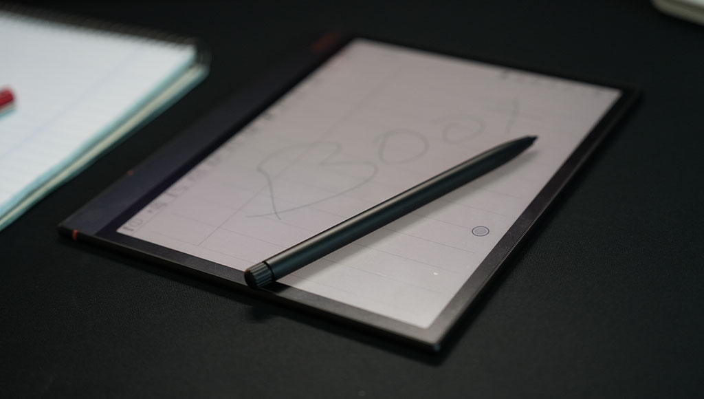 An overhead, angled shot of a Boox Note Air3 C e-reader tablet lying on a dark surface. Its screen displays a digital notepad with grid lines and the word "BOOX" lightly scribbled on it. A black stylus pen rests diagonally across the screen. Part of a spiral-bound physical notebook is visible in the upper left background.