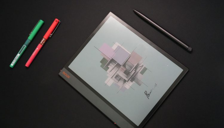 An overhead, angled shot of a Boox Note Air3 C e-reader tablet lying flat on a dark surface. Its screen displays a colorful abstract geometric design. A black stylus pen is placed diagonally above the tablet, and two colorful pens (green and red) are on the left side of the frame.
