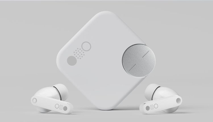 A close-up view of the CMF Buds Pro 2 in white. The square-shaped white charging case stands upright in the center, featuring a silver circular dial on its front. Two white earbuds with visible circular dots are placed on either side of the case, resting on a light grey surface.
