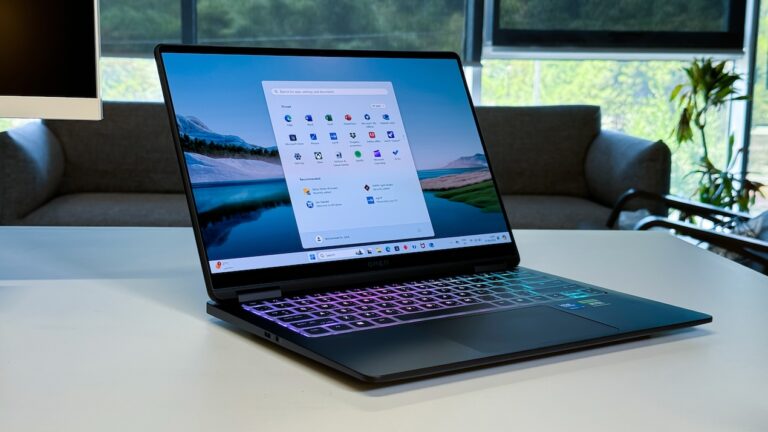 An open HP Omen Transcend 14 laptop is positioned on a white desk, with its screen displaying the Windows operating system interface and a landscape wallpaper. The laptop's keyboard is illuminated with a multi-colored (pink, purple, blue, green) RGB backlight. In the soft-focused background, a grey sofa, a window with greenery outside, and another monitor are visible.