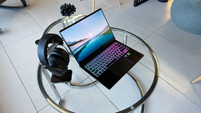 A black HP Omen Transcend 14 gaming laptop with a colorful illuminated keyboard (purple, blue, green) and a vibrant landscape wallpaper on its screen, is placed on a clear glass round table. To the left of the laptop, a black gaming headset hangs from the table's edge. A black abstract decorative object sits on the table behind the laptop. The floor is tiled in a light color, and a grey knitted pouf is visible in the background right.