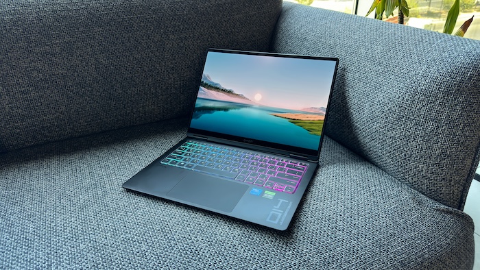 An HP Omen Transcend 14 laptop is open and resting on a textured grey sofa. The laptop's screen displays a serene landscape with a large sun reflecting on water, and its keyboard shows a vibrant RGB lighting effect in shades of blue, green, and pink.