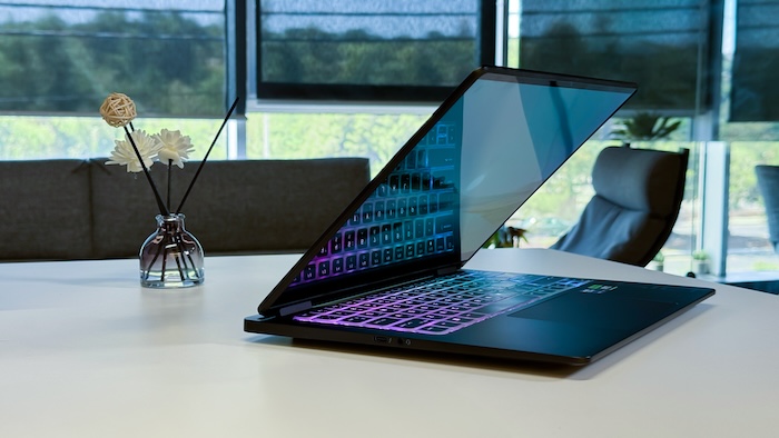 An HP Omen Transcend 14 laptop is open and angled on a white table in what appears to be a modern office setting. The laptop's keyboard is illuminated with a vibrant RGB gradient (purple, blue, green), and its screen displays colorful content. In the foreground on the left, a decorative vase with white flowers and a wicker ball on sticks adds a touch of decor.