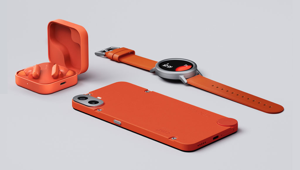A collection of CMF by Nothing orange-colored tech products displayed on a light grey surface. In the foreground, an orange CMF Phone 1 is lying face down, showcasing its back panel with visible screws and camera module. To the upper right, a CMF Watch Pro 2 with an orange band and a silver bezel is laid flat. To the upper left, an open orange CMF Buds Pro 2 charging case reveals the orange earbuds inside.