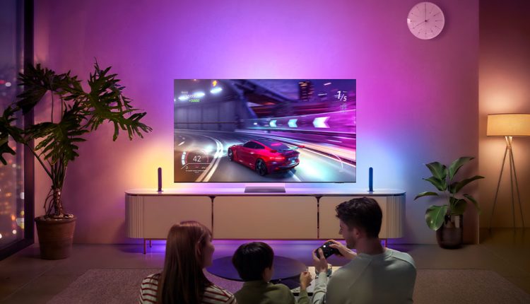 A family of three (two adults and a child) is seated on a rug, viewed from behind, in a living room brightly lit by ambient purple and pink lights from a Samsung Neo QLED 8K 2024 TV. The TV is displaying a vibrant racing video game featuring a red sports car on a track. The TV sits on a light-colored media console with two vertical light bars on either side. To the left, a large potted plant and a window showing city lights are visible. To the right, another potted plant, a floor lamp, and a white circular wall clock are present. The room has a modern and colorful aesthetic.