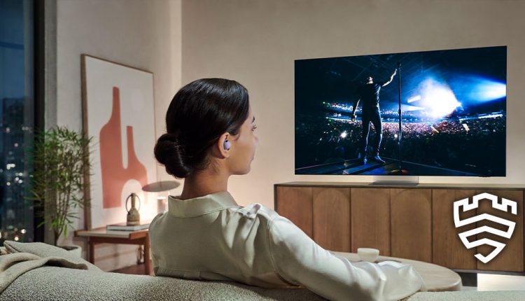 A female with dark hair styled in a bun is viewed from behind, sitting on a light-colored sofa in a modern living room at dusk or night. They are wearing a light-colored, long-sleeved top and have a light purple earbud in their right ear. The person is looking at a large, thin-bezel Samsung Neo QLED 8K 2024 TV, which is displaying a concert scene with a performer on stage illuminated by bright blue lights, facing a large crowd. The TV is placed on a wooden media console. To the left of the TV, there's a minimalist framed artwork and a plant on a side table. The city lights are visible outside a large window on the left. In the bottom right corner, a white shield icon with a stylized "S" is overlaid.