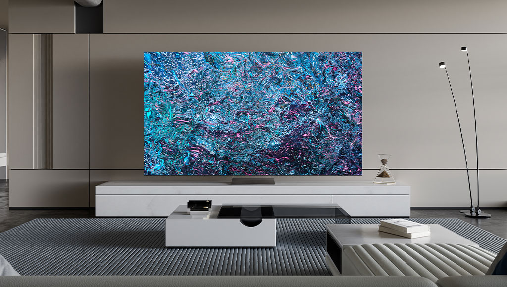 A modern living room with a large Samsung Neo QLED 8K 2024 TV mounted on a light-colored media console against a beige wall. The TV screen displays a vibrant abstract image with swirling patterns of blues, purples, and pinks. In front of the TV, there are two white rectangular coffee tables with dark glass tops. On the right coffee table, there's a small stack of white books. On the left coffee table, a glass hourglass with white sand is visible. A tall, modern black floor lamp with two light heads stands in the background on the right. The floor is covered with a striped grey rug, and a light grey sofa is partially visible in the foreground.
