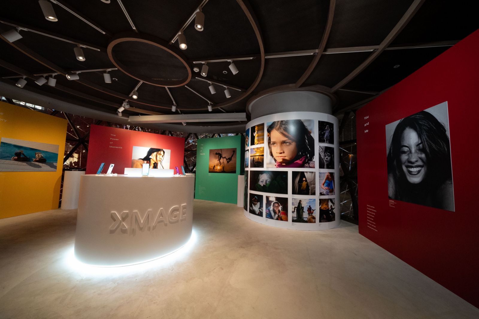 An interior view of an exhibition space featuring large photographic prints on walls and curved display panels. In the foreground, a white, illuminated circular counter displays the "XMAGE" logo. Various colored walls (yellow, red, green) are visible, showcasing diverse photography styles, with some Huawei phones on display on pedestals. The ceiling has spotlights illuminating the space.