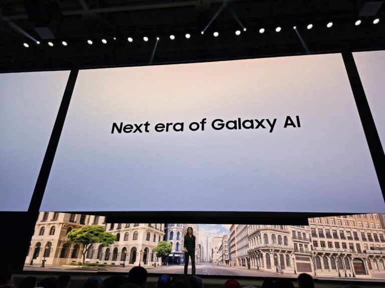 A person stands on a large stage in front of a massive screen displaying the phrase "Next era of Galaxy AI" during an official Samsung event unveiling the next generation of AI technology. A virtual backdrop of a European-style street is shown at the bottom of the screen, with an audience visible in the foreground.