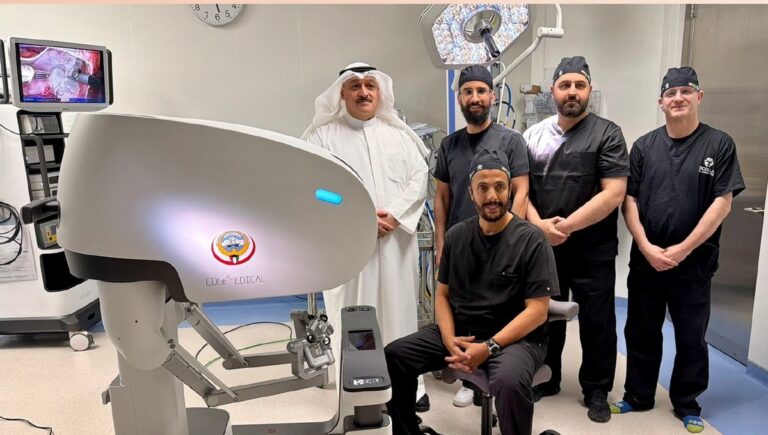 صورة جماعية داخل غرفة العمليات تظهر فريقاً طبياً يقف بجانب جهاز "Edge Medical" للجراحة الروبوتية الذي يحمل شعار دولة الكويت. يظهر في الصورة جراح يجلس أمام وحدة التحكم بالروبوت، ويقف بجانبه مسؤول يرتدي الزي الخليجي التقليدي (الدشداشة والغترة)، بالإضافة إلى ثلاثة جراحين يرتدون زي العمليات، مع ظهور شاشة تعرض مجريات الجراحة في الخلفية